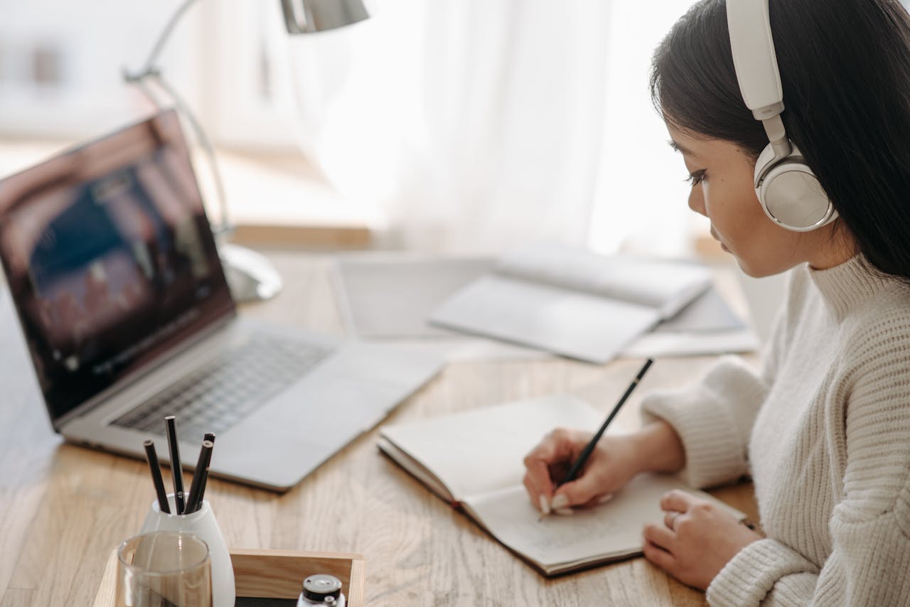 Asian woman focused on study, writing notes during online session at home with laptop and headphones.