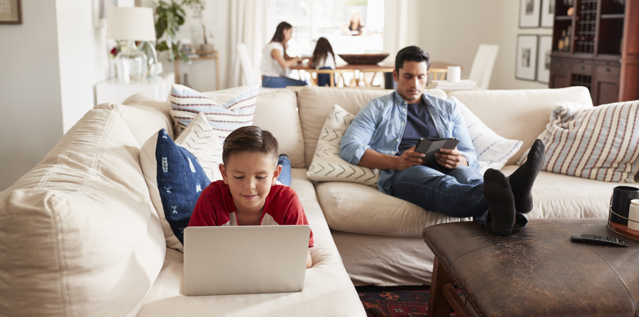 pre teen boy lying on sofa using laptop, dad sitting with tablet, mum and sister in the background