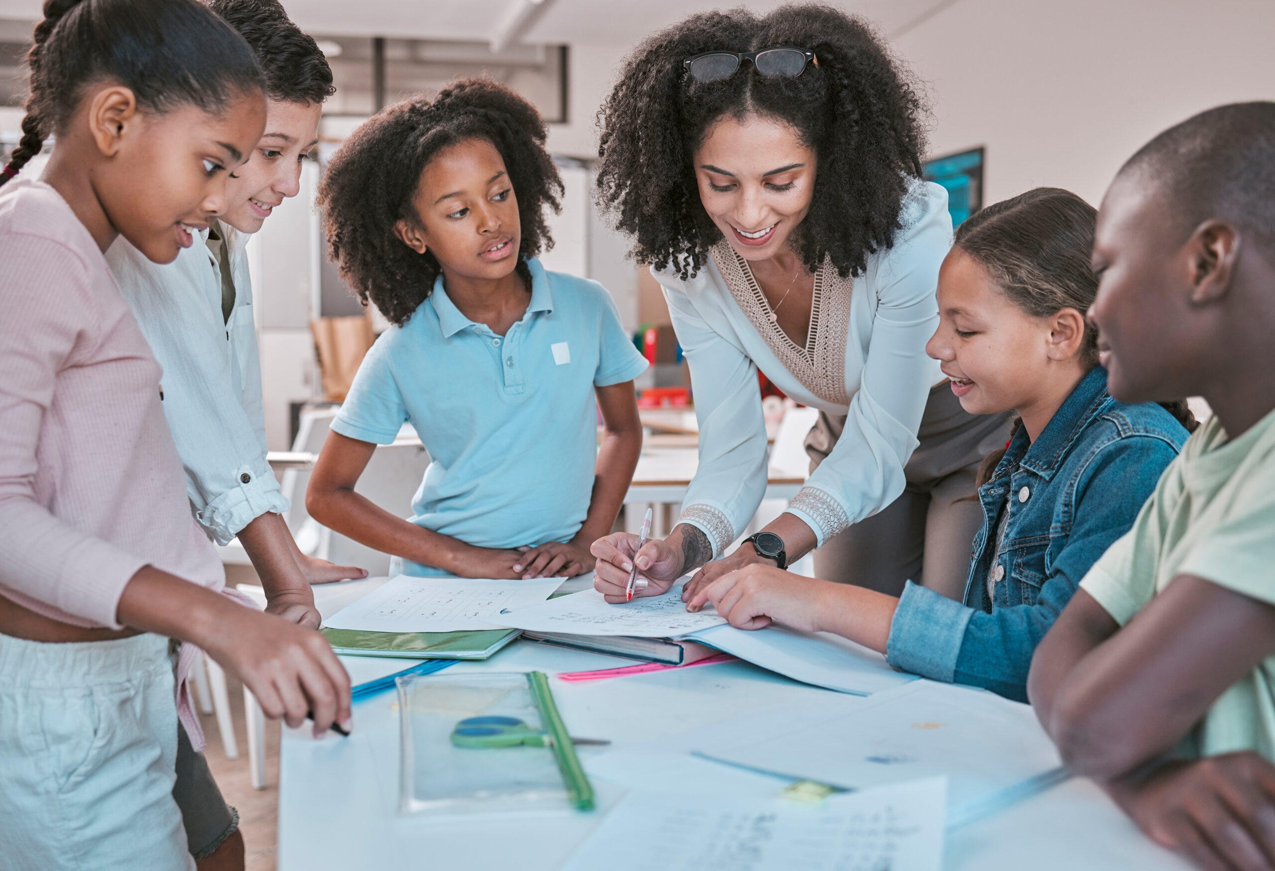 female teacher in classroom with students, helping learner with homework and writing in book. children's education at school, educator reading kids notebook and group learning together for assessment