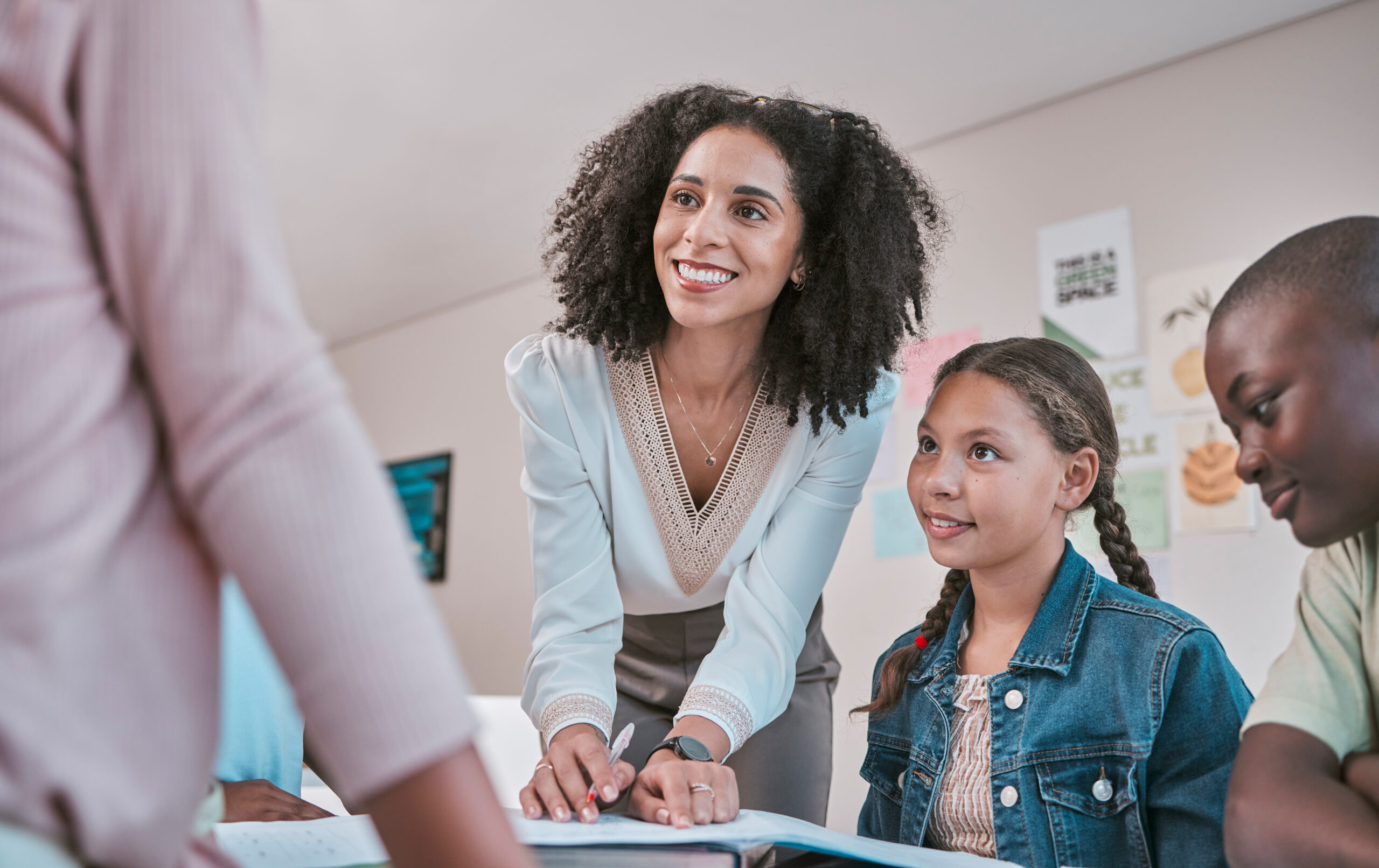 happy teacher in classroom with children, helping students with homework and writing in book. education in school, female educator reading kids notebook and group learning together for assessment