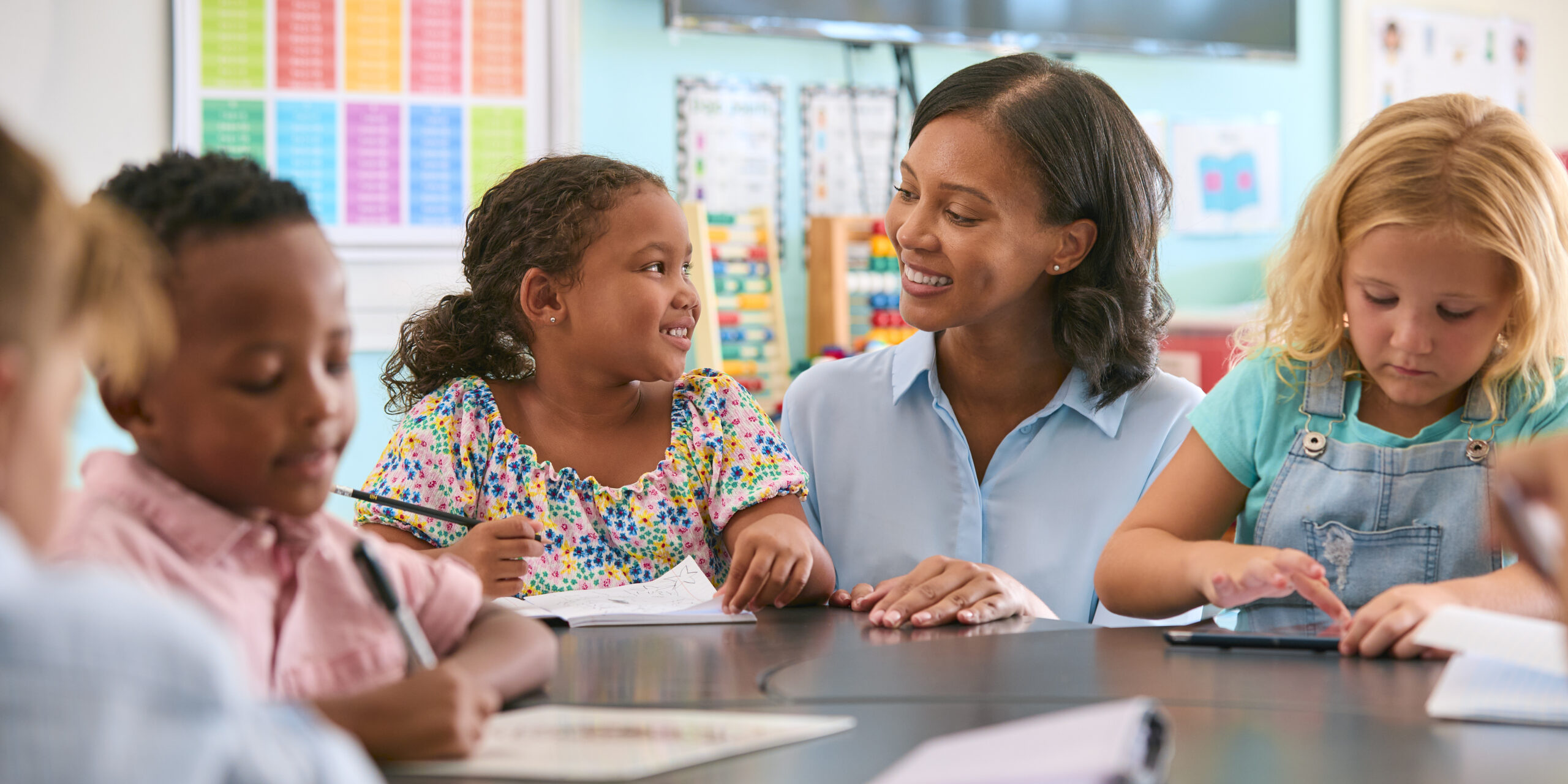 female primary or elementary school teacher helping students at desk in multi cultural class
