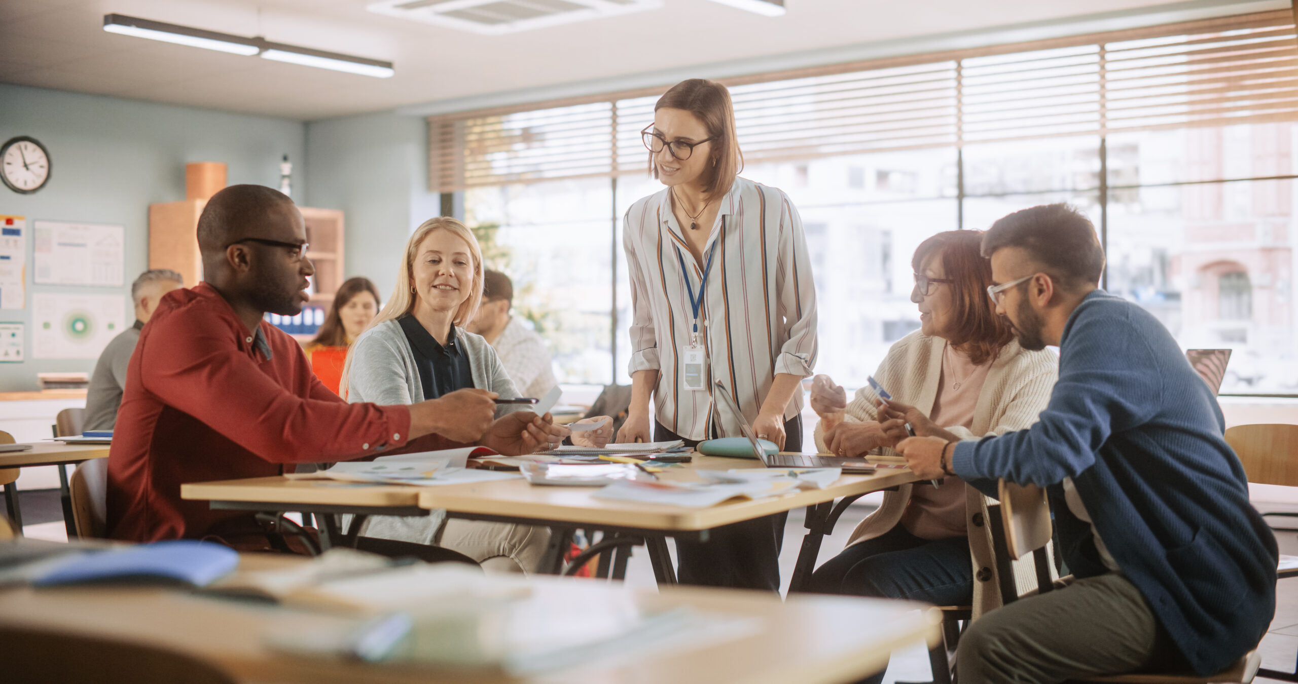 adult education center: adult students learning in classroom, working as two teams on a verbal education game. positive female teacher giving advice, answering questions, and motivating students