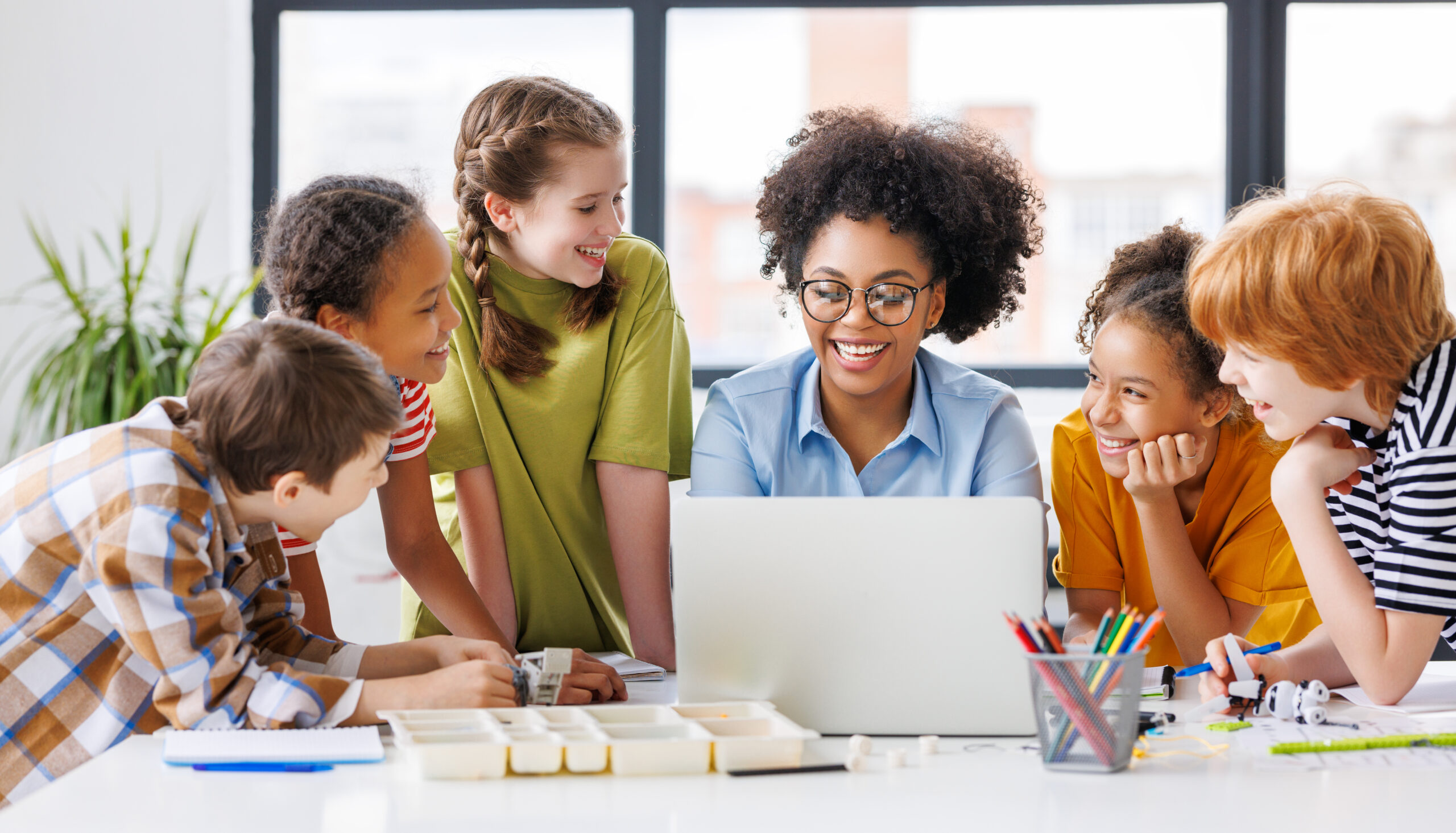 cheerful schoolkids with teacher looking at laptop screen and study the scheme of assembly of robots during collective school work