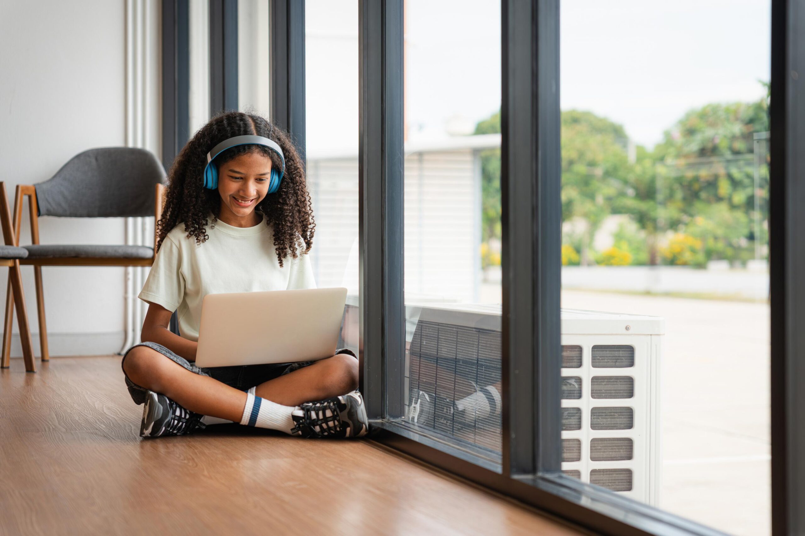 How to Choose a Flexible, Accredited K-12 Virtual School That Fits Your Family’s Needs in 2026-2027 a teenage girl with curly hair and headphones smiles while using a laptop, sitting on the floor by a window, portrait of a relaxed student enjoying entertainment with a laptop and headphones
